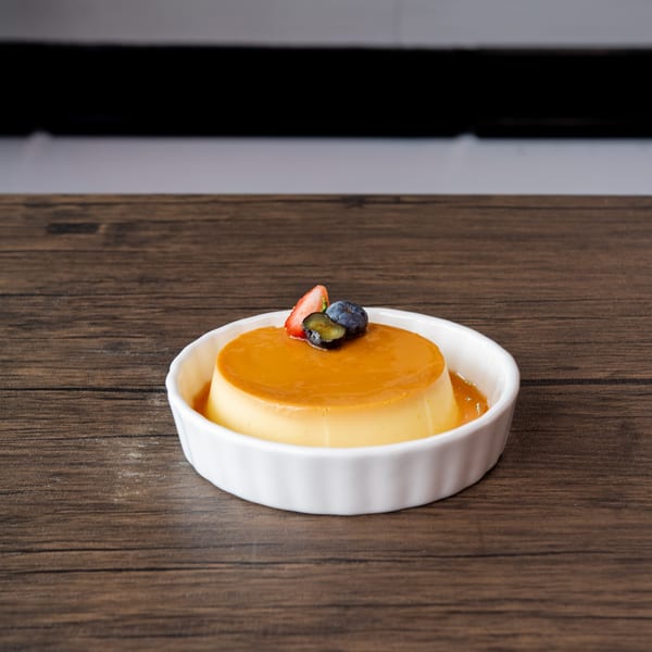 A creamy flan dessert with caramel sauce, topped with a strawberry and blueberries in a white dish.
