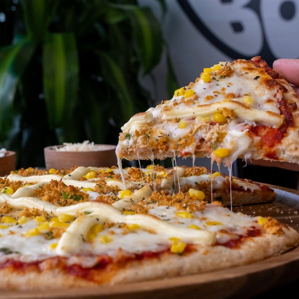 Hand lifting a cheesy slice of pizza with chicken and corn from a wooden plate.