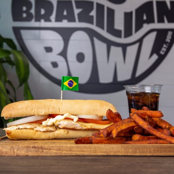 A Brazilian chicken sandwich with sweet potato fries and a drink on a wooden board.
