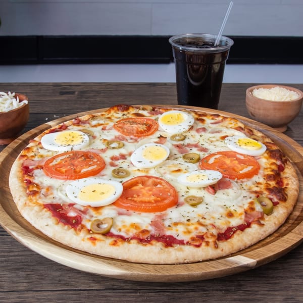 A pizza with tomatoes, olives, ham, and boiled eggs, served with a drink and cheese.