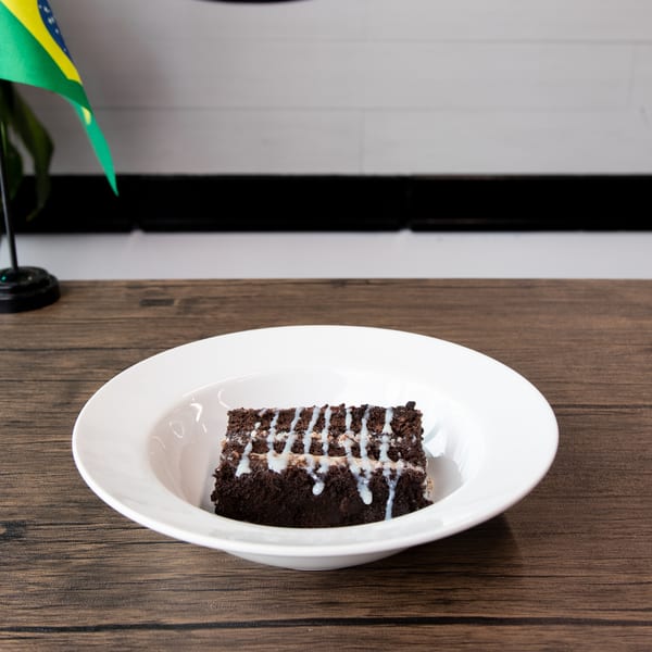 A slice of chocolate cake with white icing drizzle in a white bowl on a wooden table.