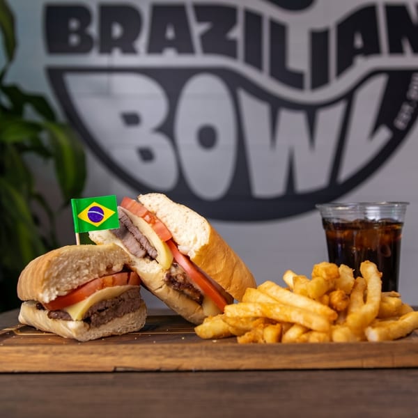 Two Brazilian steak sandwiches with cheese and tomato, fries, and a soda on a wooden board.